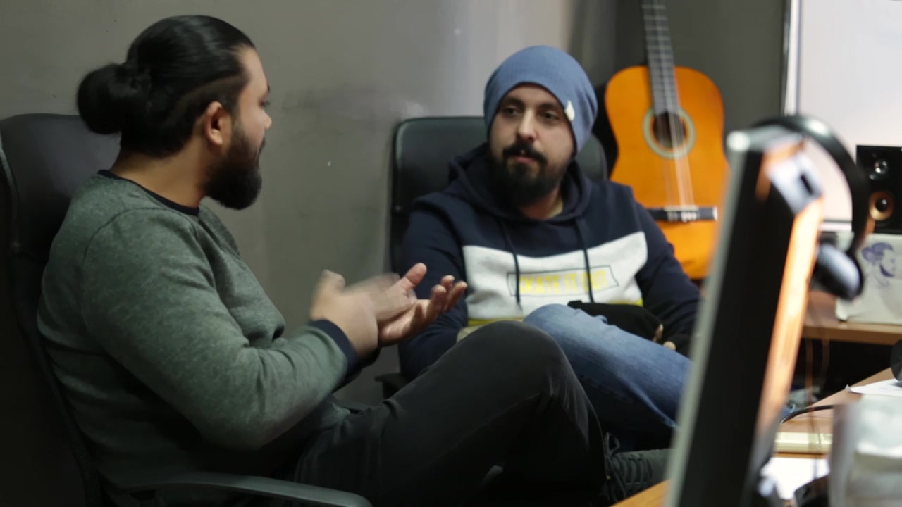 Ofiste müzik üzerine sohbet eden, şarkı dinleyip elleriyle ritme eşlik eden genç erkekler, fonda gitar.