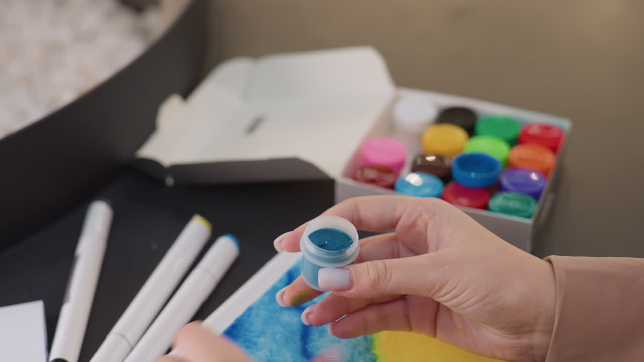 Mujer abriendo un pequeño bote de pintura azul; mujer abriendo con cuidado su bote de pintura azul sobre la mesa; persona desatornillando suavemente un bote de pintura azul entre materiales de arte esparcidos sobre una mesa