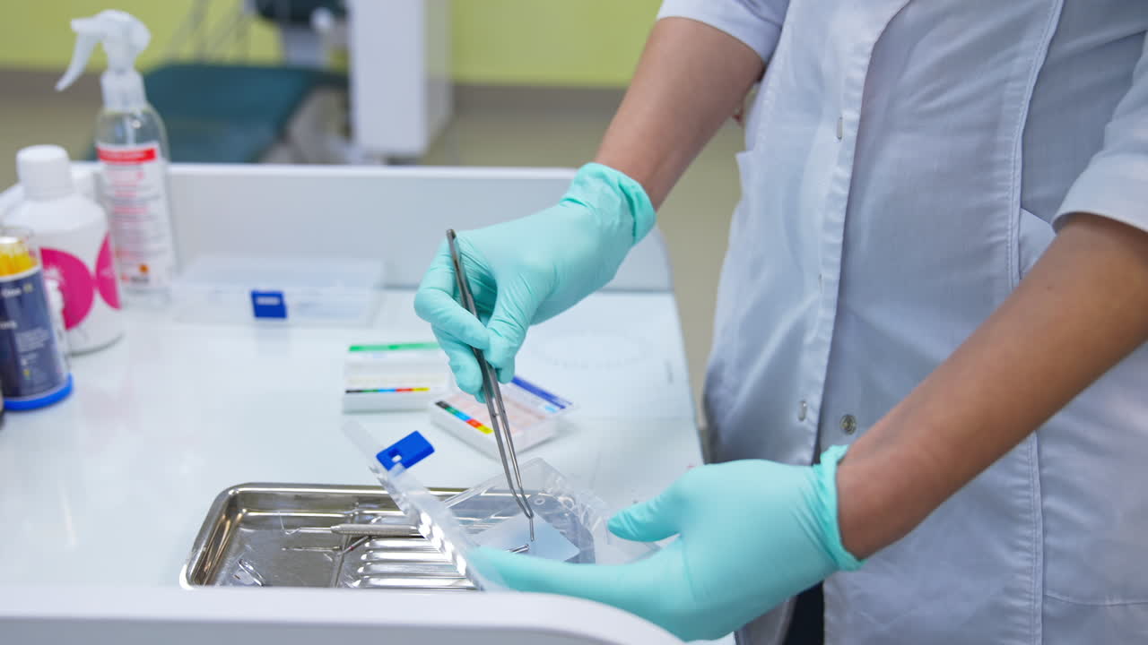 Person in white coat and blue latex gloves holds metal forceps. Metal nozzles are taken from a plastic box and put into the steel tray on the drawer. Close up.