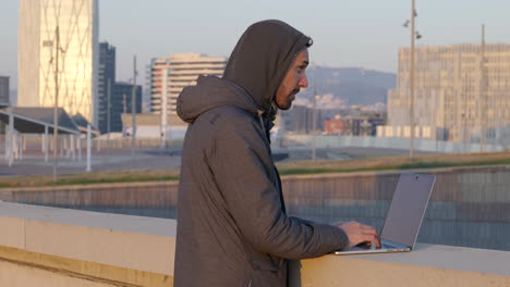 Wintry-dressed-male-hacker-aware-of-surroundings-typing-on-laptop-at-Barcelona-waterfront-business-district