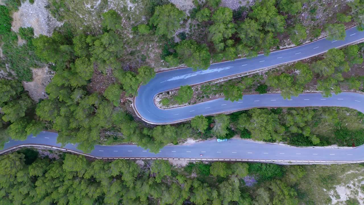 Top-down aerial view of winding mountain road near Mirador de Es Colomer, Mallorca. Serpentine bends cut through lush forest in a scenic Mediterranean landscape