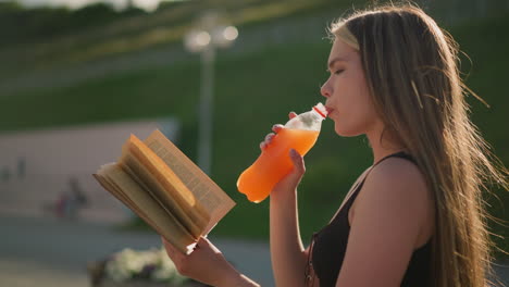 side view of a woman seated outdoors holding a book in one hand while sipping from a juice bottle in the other, she continues reading, with blurred background featuring lamp pole and greenery hill