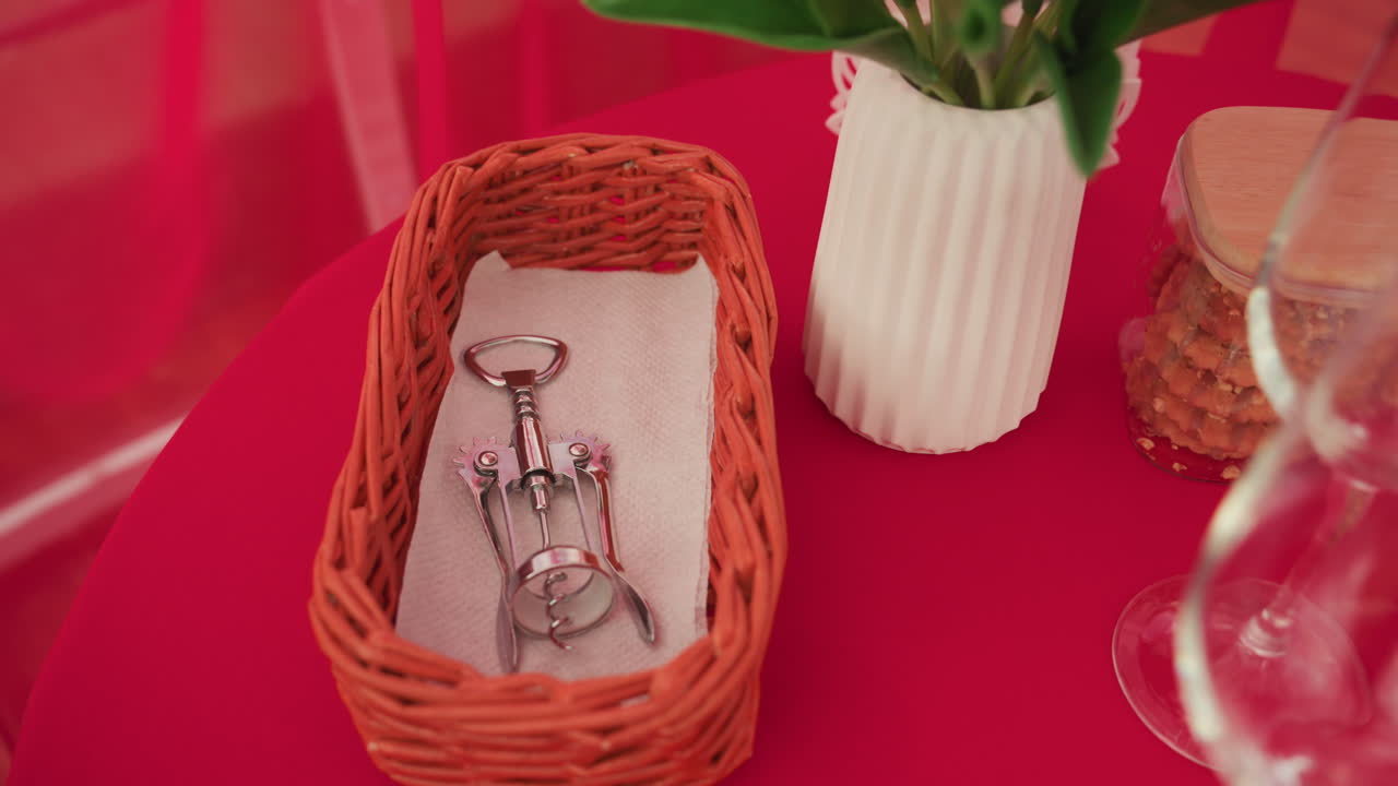 Tavolo rosso con accessori per il vino e cesto di vimini contenente cavatappi, tulipani in vaso di ceramica, bicchieri e barattolo di tappi di sughero, illuminazione bokeh soffusa che crea un'atmosfera intima, accogliente e romantica per un appuntamento di coppia