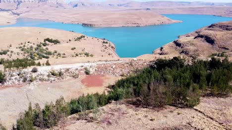 Toma-Aérea-Que-Asciende-Por-Encima-De-La-Línea-De-árboles-Para-Revelar-Una-Vista-Panorámica-De-La-Presa-Driekloof,-Sus-Aguas-Azules-Añaden-Color-Al-Paisaje-Seco-Y-árido-De-Invierno-Que-Rodea-La-Presa,-Estado-Libre,-Sudáfrica