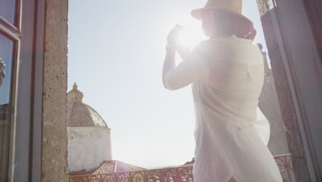 happy woman using smartphone taking photo enjoying sharing summer vacation travel experience photographing beautiful scenic view on balcony wearing hat