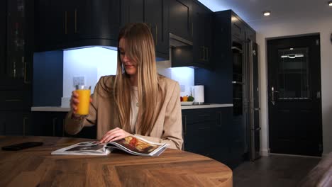 female model in a smart jacket drinking from a glass of orange juice reading a magazine at a kitchen table in a modern kitchen