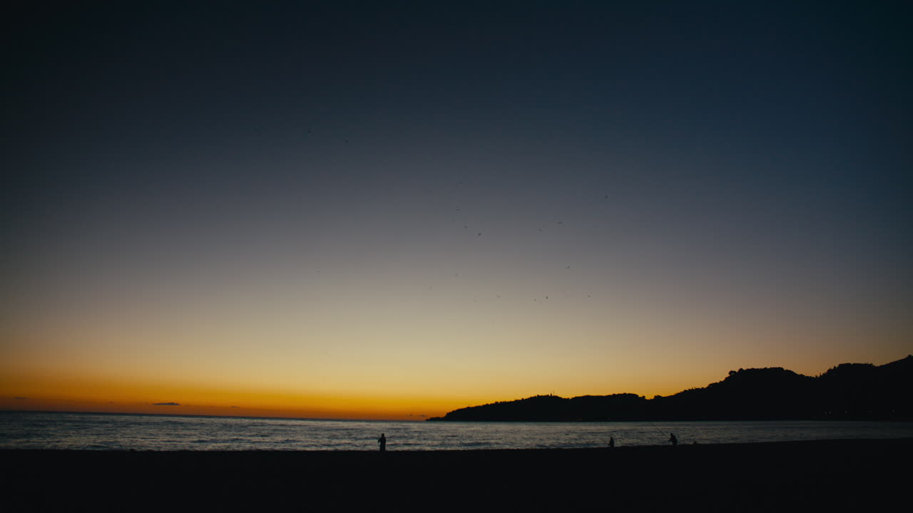 Wide shot of several fishermen silhouetted against a vivid coastal sunset, as seabirds scatter across the colorful sky. The scene captures a serene moment of solitude and nature by the sea