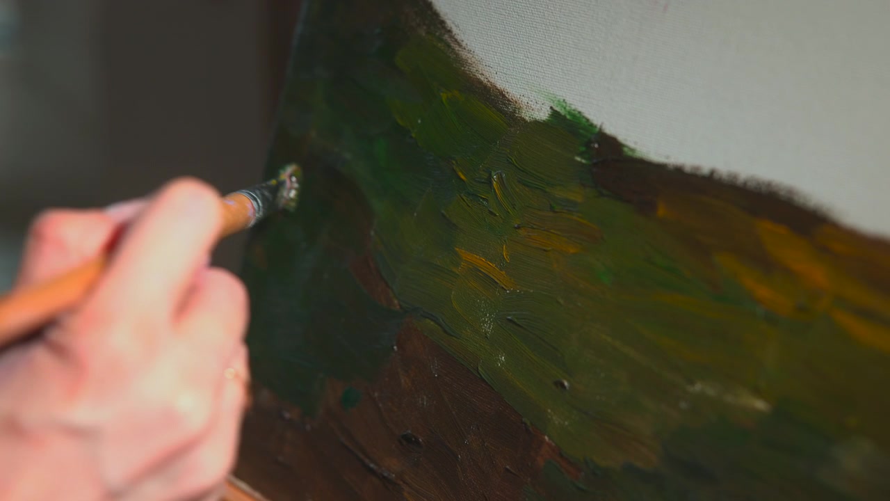 Vista ravvicinata di una mano che usa un pennello per applicare pittura su una superficie testurizzata verde e ambra, con illuminazione soffusa, messa a fuoco ridotta e movimento visibile del pennello.