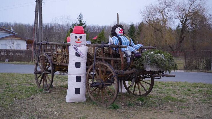 A rural wooden cart decorated with a snowman and a clown figure, filled with hay and greenery