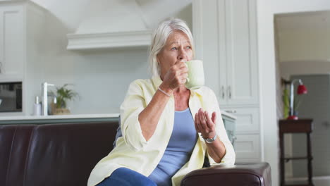 Happy-caucasian-senior-woman-drinking-coffee-and-smiling-on-sofa-in-sunny-living-room,-slow-motion