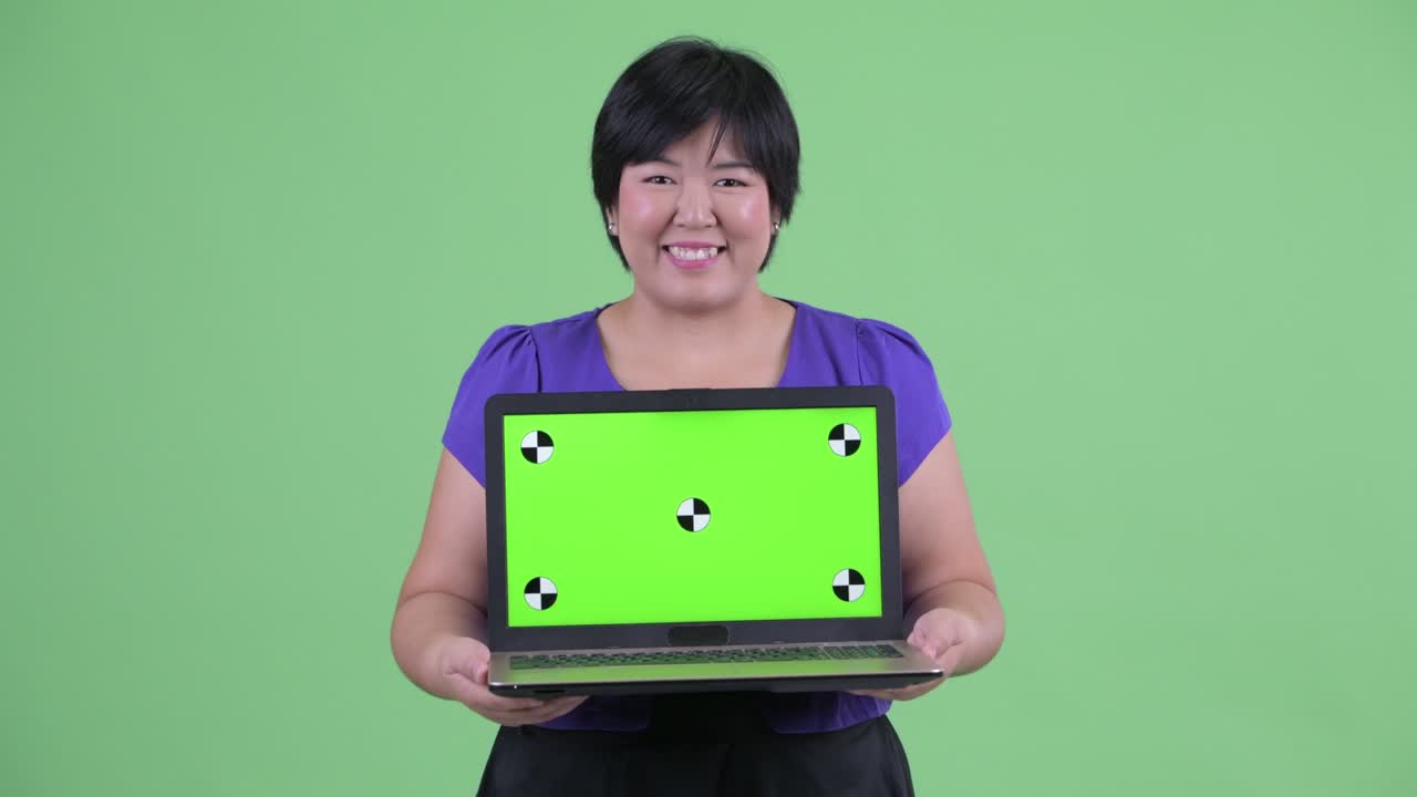 초록색 배경에 미소 짓는 동안 컴퓨터에서 크로마 키를 보여주는 짧은 헤어 스타일의 과체중 아시아 여성의 초상화