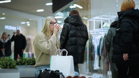 young lady sipping coffee, looking thoughtful, with decorative flower close by, background features a clothing store with shoppers walking by in the mall, creating a busy atmosphere