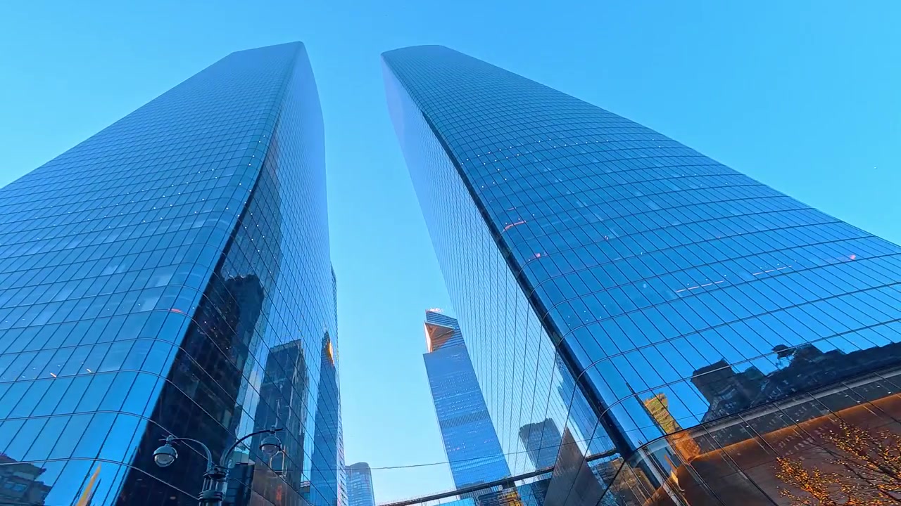 Glasskyskrabere, der strækker sig mod den blå himmel ved Hudson Yards i New York City. Moderne spejlfacader reflekterer sollys fra et lavt perspektiv nær et overhængende trafiklys