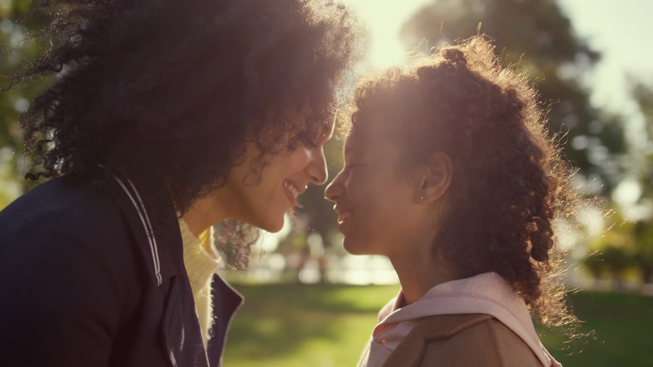 Smiling parent touching noses with cute daughter in golden sunlight portrait.