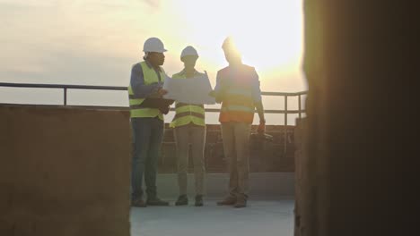 caucasian woman boss with laptop computer standing on the sunset on the roof with two male multiethnic builders.