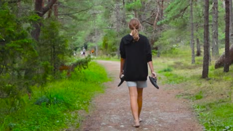 girl with a ponytail who is holding flip flops walking down on a rural forest road or track during summer time in slow motion. nature and trees surrounding the road. girl has shorts and a black shirt.