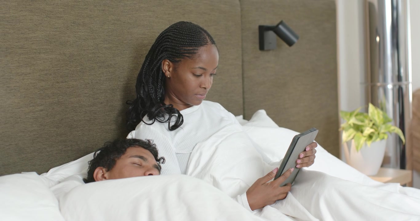 African couple lying in bed while man sleeping and woman holding tablet, reading to unwind