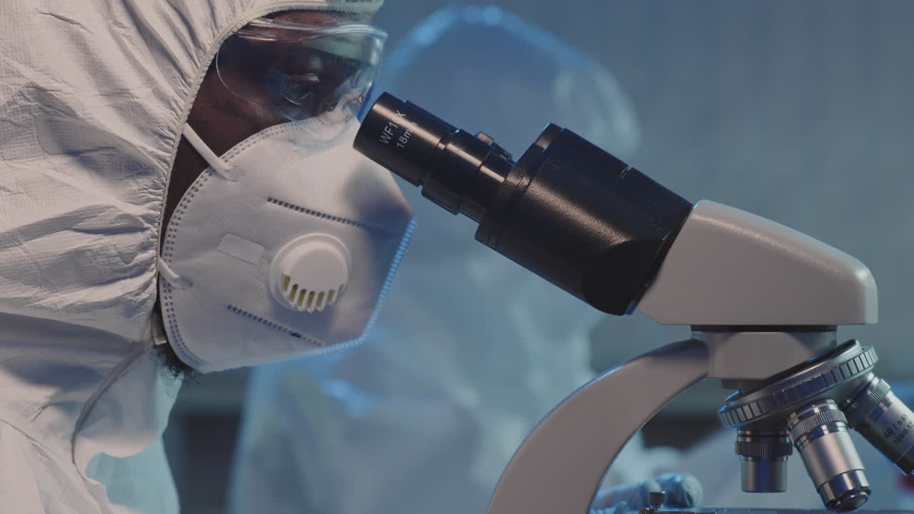 Scientist in Coveralls and Face Mask Studying Sample under Microscope