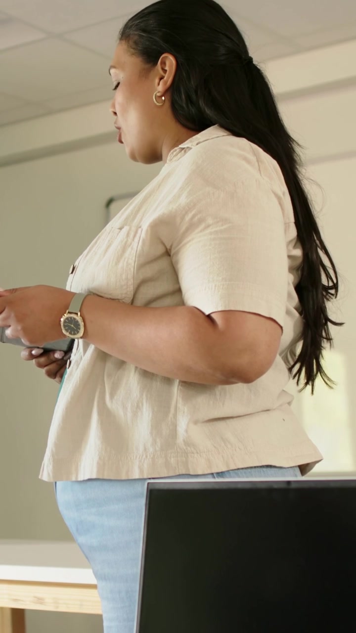 Video verticale: Una presentatrice con una camicetta beige guarda il telefono mentre tiene un briefing in aula.