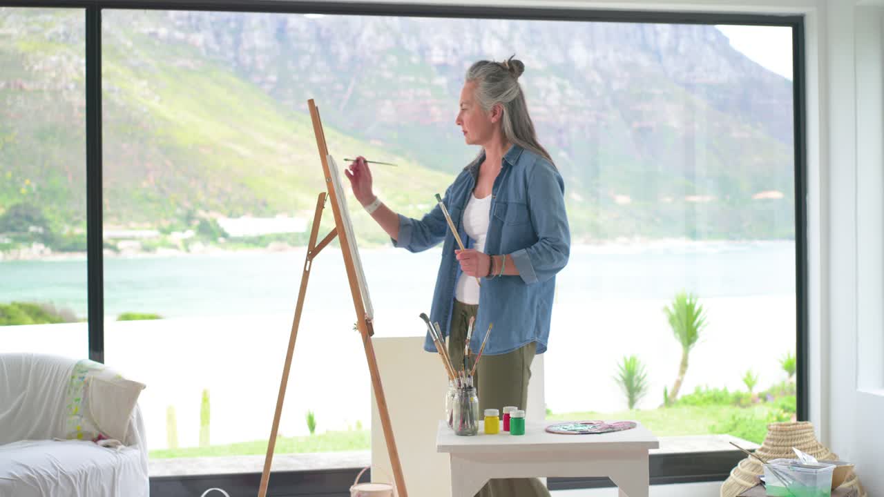 Reife Frau in Jeans im Studio, die sich von einem Seeblick inspirieren lässt, wählt Pinsel aus, mischt Farbe und malt