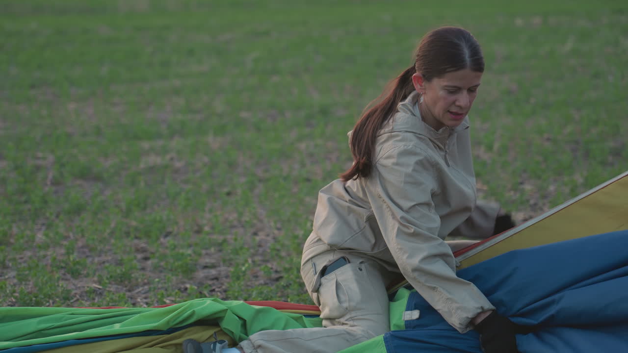 woman squatting on open green field folding multicolour hot air balloon fabric into neat bundle along durable ropes while preparing for transport under soft evening light