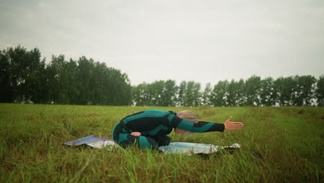 Side view of woman in green and black suit seated on yoga mat with legs crossed, hands raised above her head, practicing yoga in a grassy field, with yellow boots visible in the background