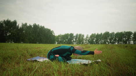 side view of woman in green and black suit seated on yoga mat with legs crossed, hands raised above her head, practicing yoga in a grassy field, with yellow boots visible in the background