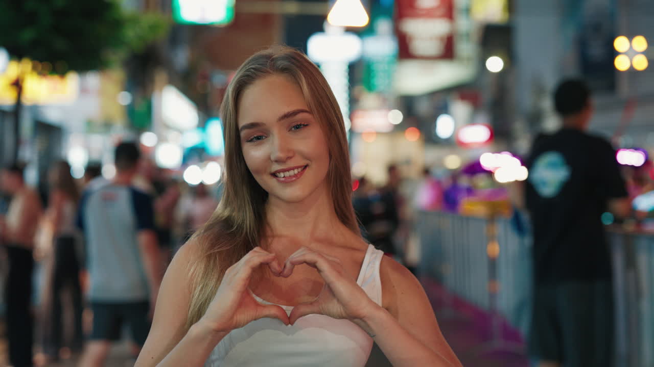 Happy young woman making a heart shape with hands in a city at night