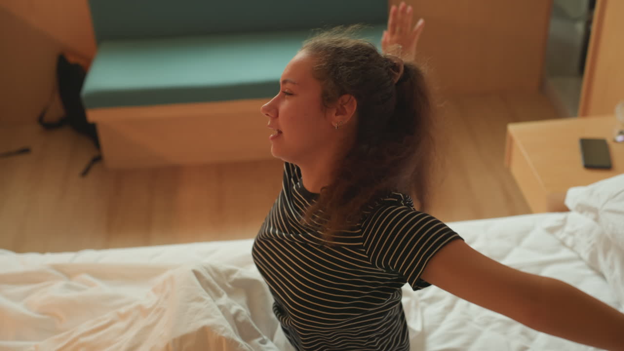 Donna di colore seduta sul letto d'hotel, con top a righe e capelli ricci, luce solare calda che attraversa il pavimento in legno, panca e comodino con telefono e bicchiere, atmosfera tranquilla e contemplativa, ospite solitario che riflette prima dell'inizio della giornata.