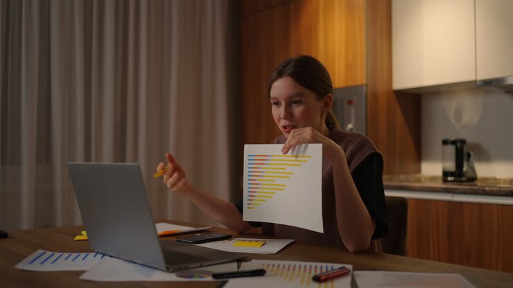 Remote work A young woman shows a graph in a laptop camera while sitting at home in the kitchen conducting an online report. Video call and conference with demonstration of documents and schedules