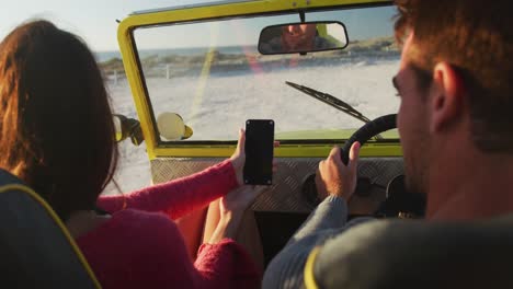 Feliz-Pareja-Caucásica-Sentada-En-Un-Buggy-De-Playa-Junto-Al-Mar-Usando-Un-Teléfono-Inteligente