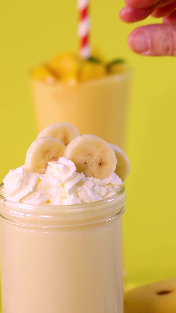 Una mano posiziona fette di banana su un cremoso milkshake alla banana con panna montata, sfondo giallo brillante, primo piano.