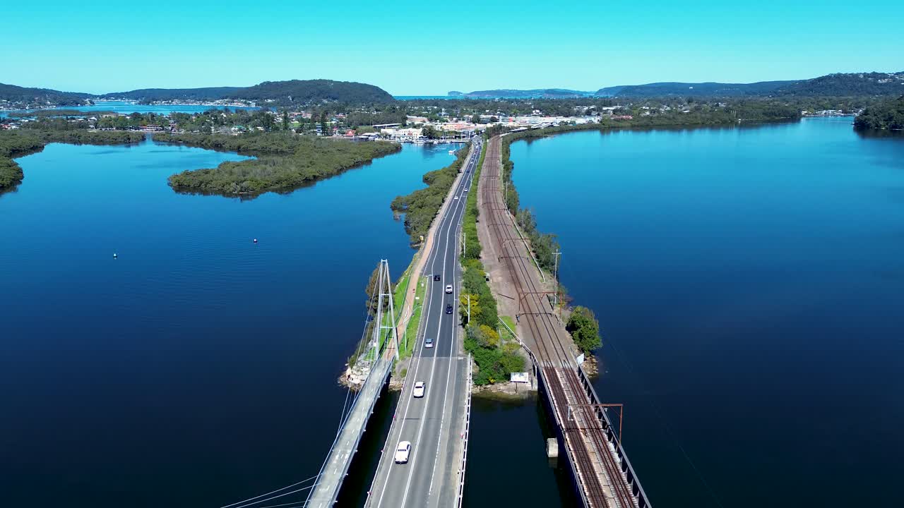 Drone aerial landscape of cars driving along main road highway bridge into Woy Woy town suburbs with train railway line running alongside Central Coast Australia travel transport infrastructure