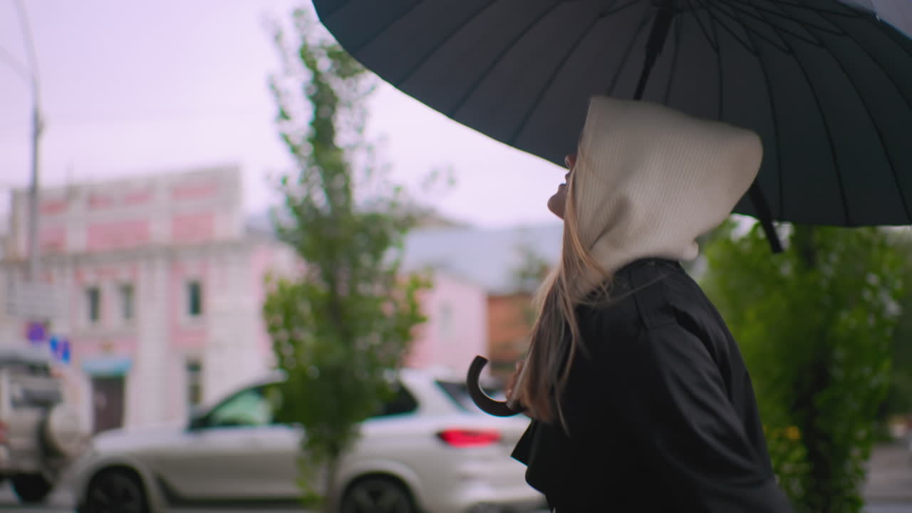 Maiden wearing black coat and white hood running with large black umbrella on city street during overcast weather, cars and buildings visible in background creating moody urban atmosphere of rainy day commute