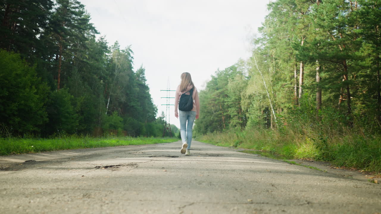 Back view of young woman walking along quiet tarred road surrounded by lush forest, carrying backpack with relaxed pace on peaceful day under overcast sky, reflecting solitude