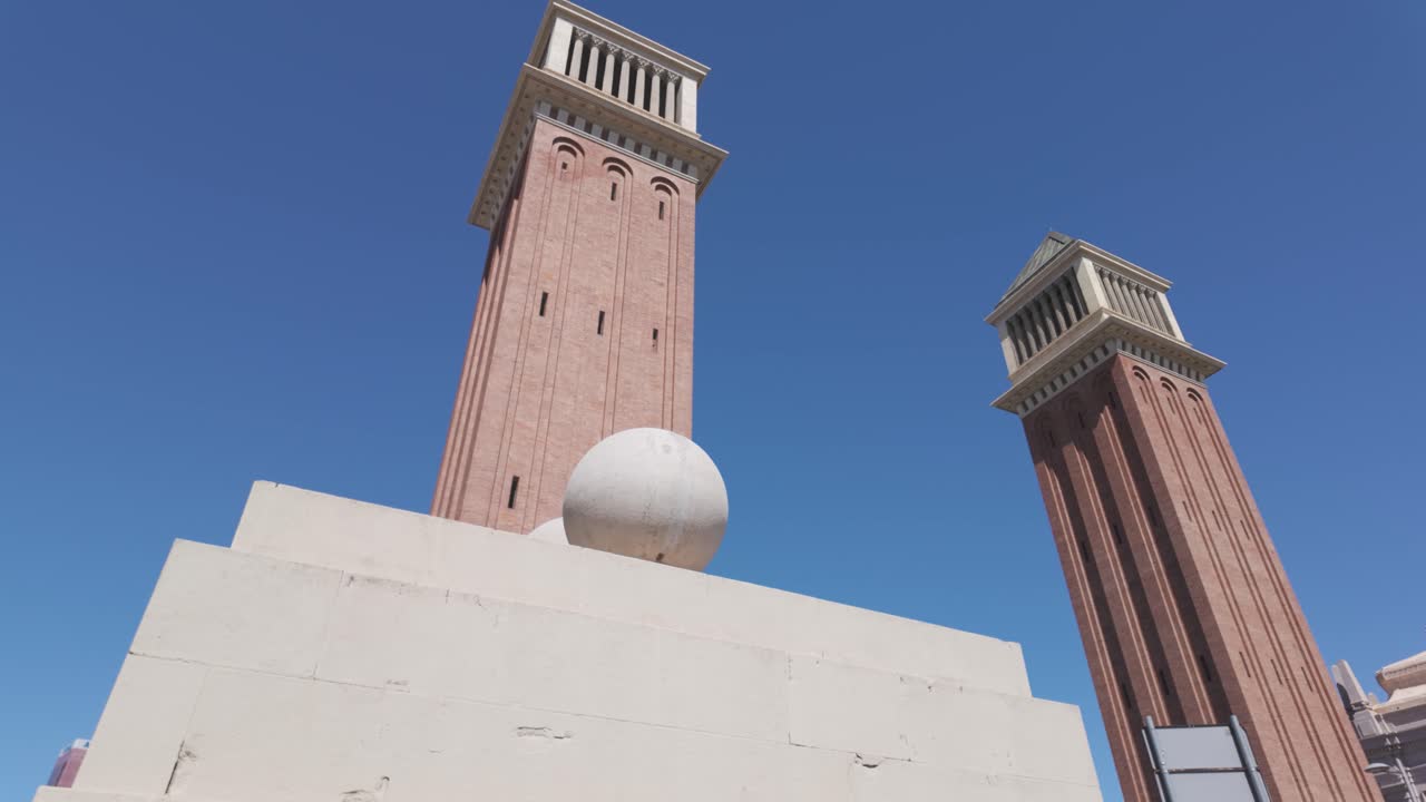 바르셀로나의 밝은 파란 하늘을 배경으로 두 개의 상징적인 베네치아 타워가 건축적 우아함과 역사적 의미를 보여줍니다.