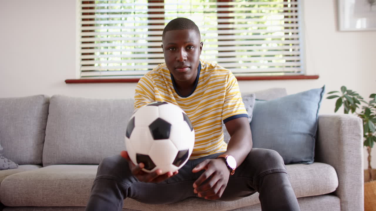 Holding soccer ball, african american man sitting on couch and looking at camera, at home