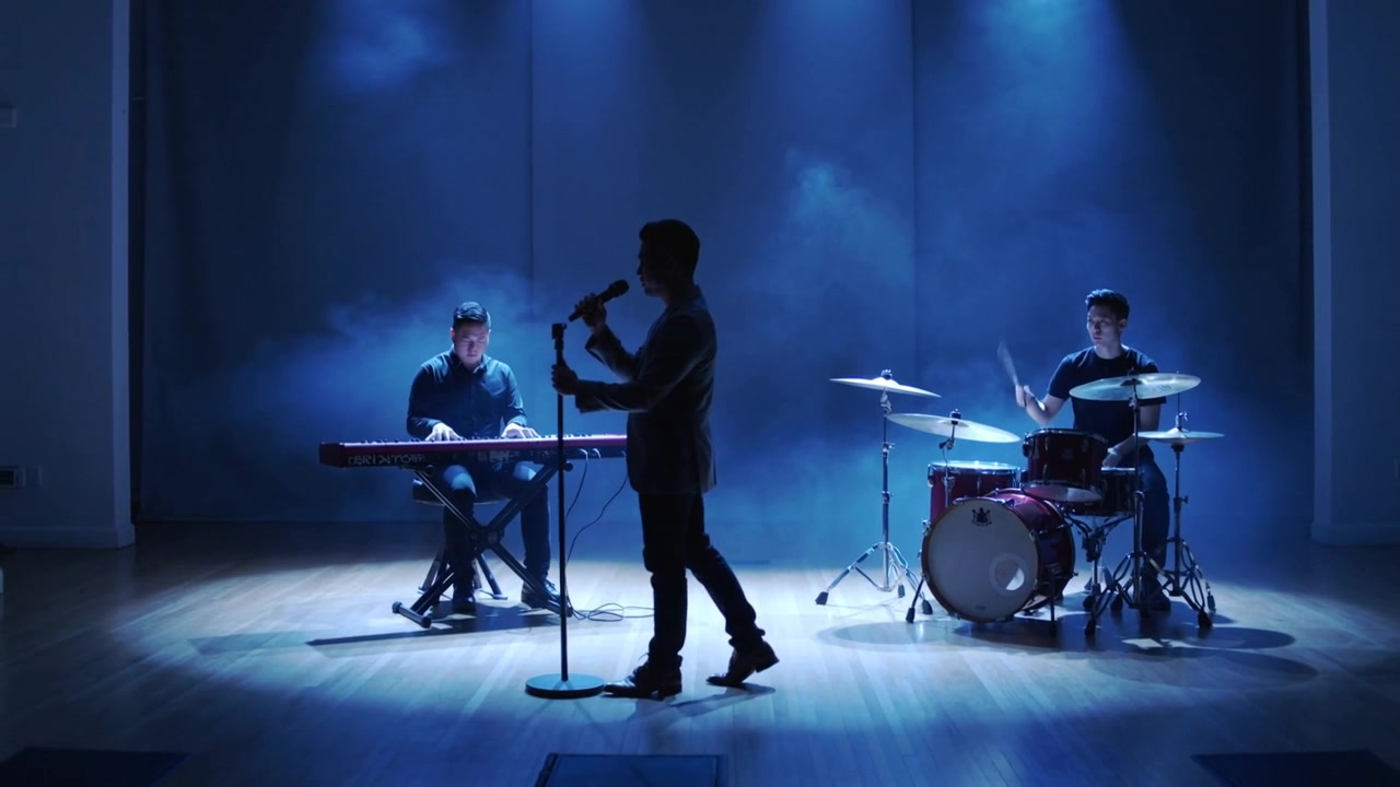 Cantante che canta e aggiusta la presa del microfono in studio, mentre i riflettori illuminano una band in abiti scuri che suona tastiere e batteria.