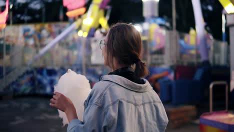 Tracking-footage-of-a-young-blonde-girl-walking-in-the-amusement-park,-taking-white-candy-cotton.-Looking-over-her-shoulder-and