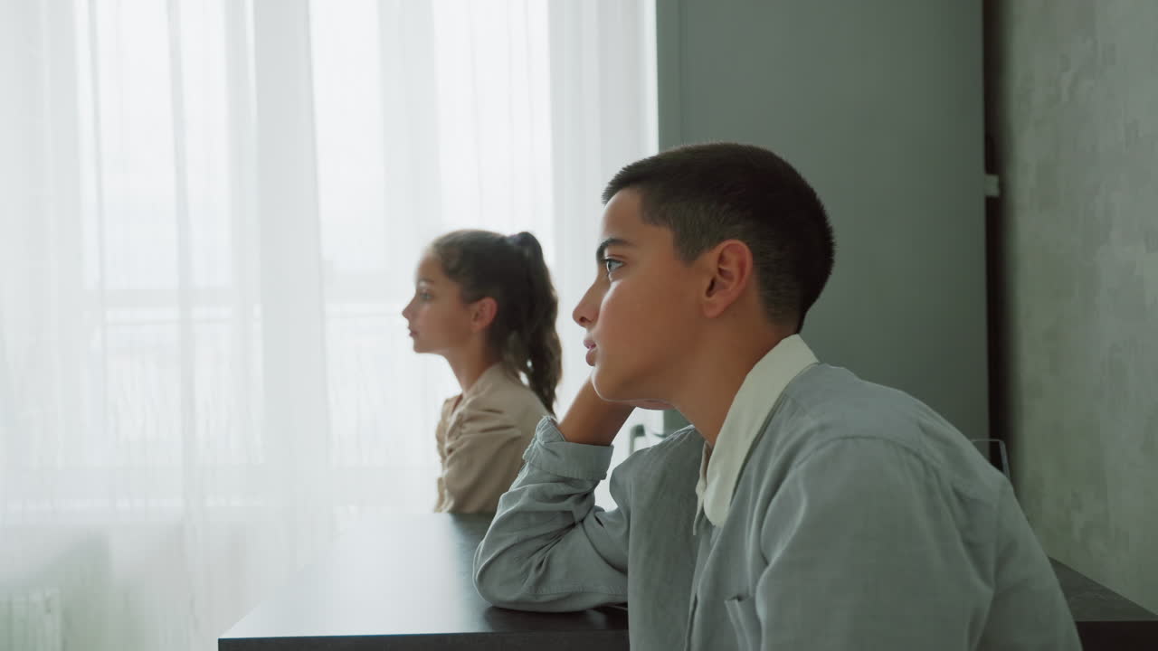 Kids in school uniforms sitting quietly at table near soft white curtain in bright morning light with thoughtful expressions indoors during calm home routine before leaving for school