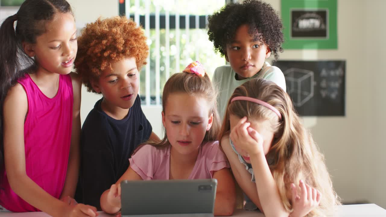 Bambini multietnici riuniti intorno a un tablet a scuola, una bambina seduta che tocca per giocare, i coetanei che si sporgono per vedere