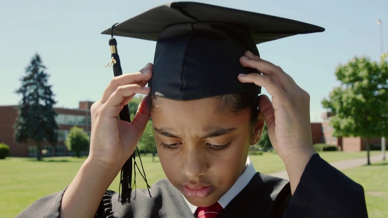 Noticing mortarboard slipping student in black gown adjusting cap on campus lawn securing tassel