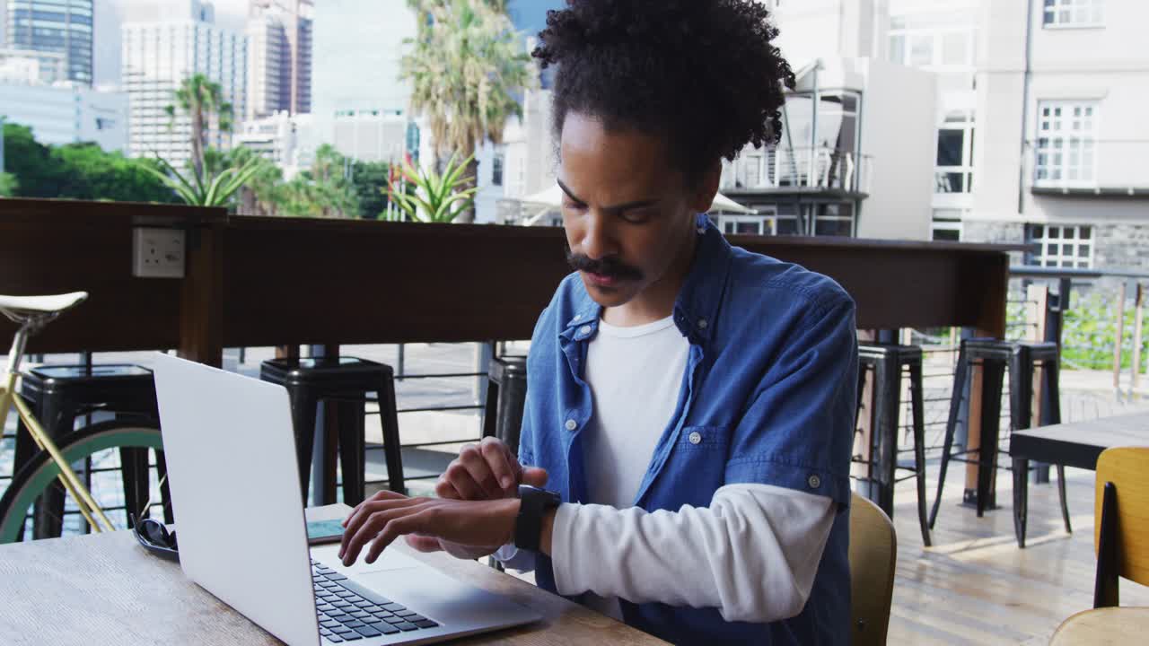 hombre de raza mixta con bigote sentado en la mesa fuera del café usando una computadora portátil y un reloj inteligente