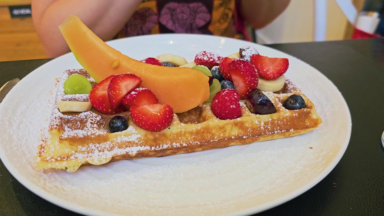 Waffle croccante guarnito con fragole fresche, mirtilli, lamponi, banana, uva, melone e una spolverata di zucchero a velo, in un ambiente da colazione di un ristorante informale con luce calda