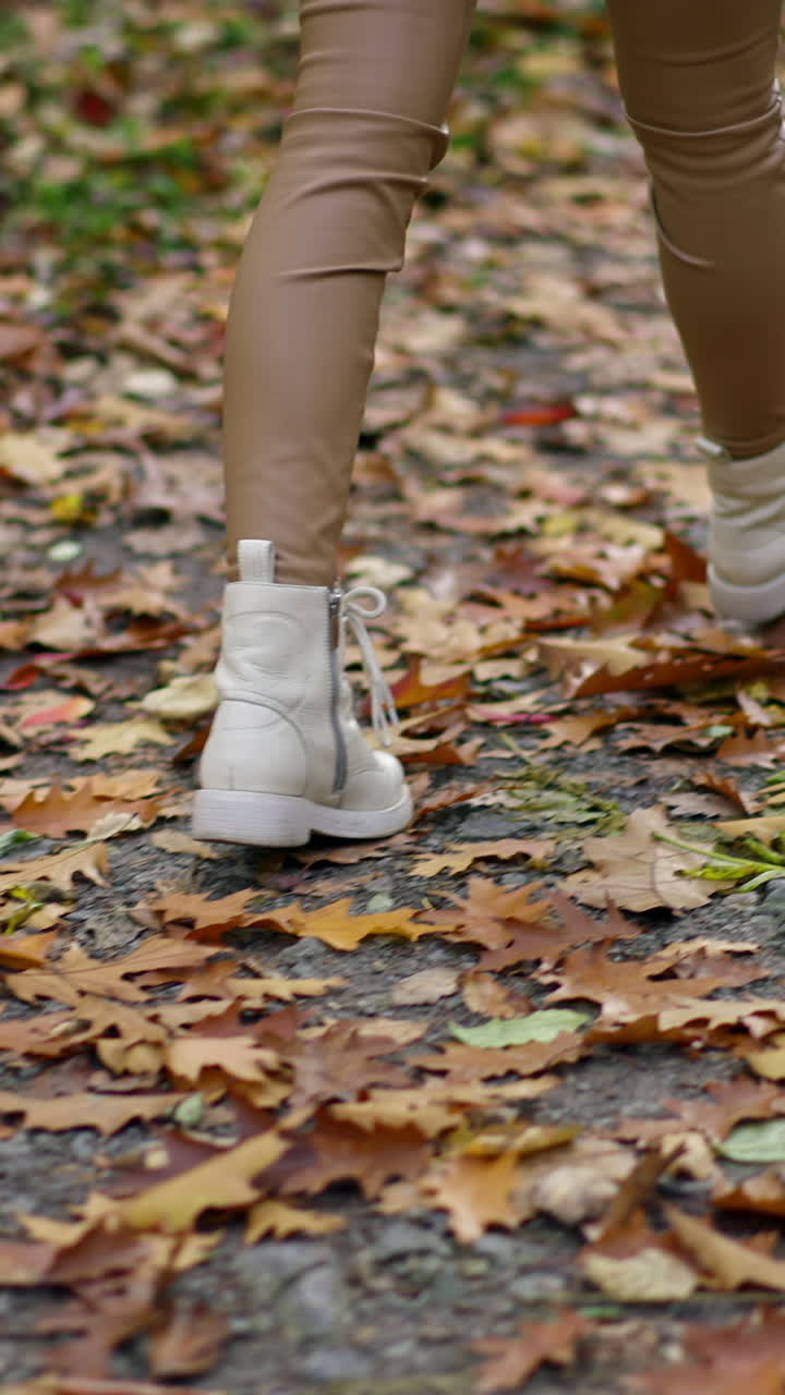 Female in tight beige leggings and white boots walks quickly by the path in the park. Unrecognized lady taking a stroll on autumn day.