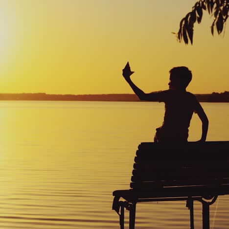a little boy is sitting on the bench and playing with a paper boat on the background of evening landscape by the lake in the summer. sunset