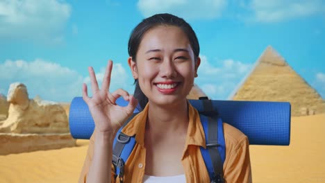 close up of asian female hiker with mountaineering backpack smiling and showing okay gesture to camera while traveling in pyramid of giza