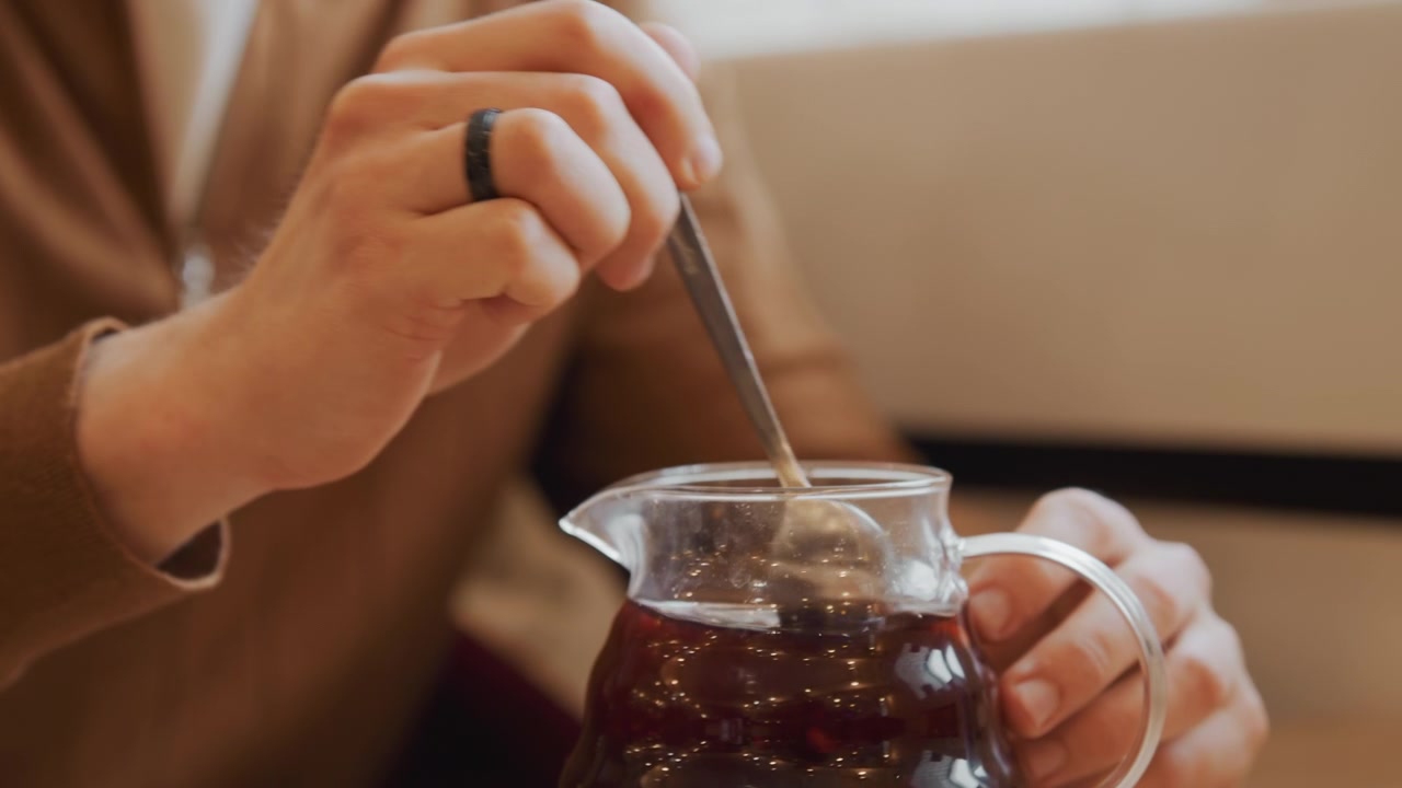 Primo piano di un uomo che mescola un liquido scuro in una brocca di vetro con entrambe le mani, indossando abiti marroni e un anello nero. La scena presenta un'illuminazione soffusa su uno sfondo di tavolo in legno, concentrandosi esclusivamente sulla mano.