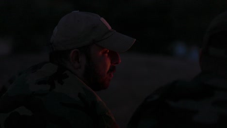 close-up of a bearded man's face in camouflage clothes and cap at night lit by campfire flames, he tells a scary story to his friends, night camp, hunting