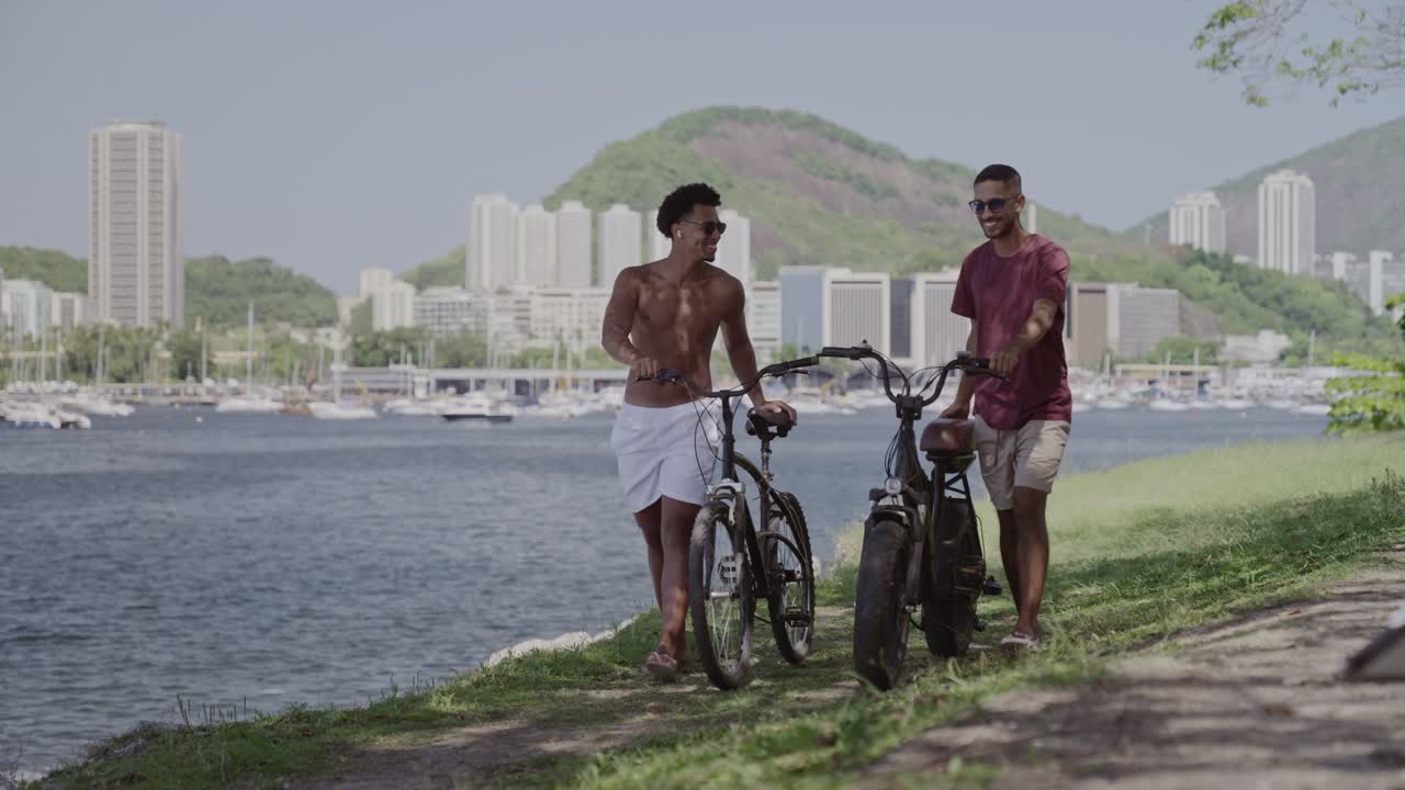 Two Men Walking Bicycles Along a Scenic Waterfront with a Cityscape Background
